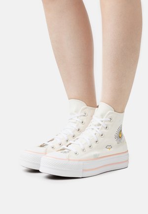 Baskets montantes blanches avec des motifs de soleil, nuage et éclair portées aux pieds sur un fond uni.