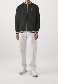 Tmavě zelená bomber bunda s bílými pruhy na manžetách a lemu, zipsové zapínání, vyšívané logo na hrudi, doplněná o světle barevné kalhoty a tenisky.