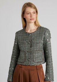 Grüne Tweedjacke mit strukturiertem Design, Rundhalsausschnitt, Fronttaschen und Knopfverschluss. Abgerundet mit braunen Paspelakzenten.