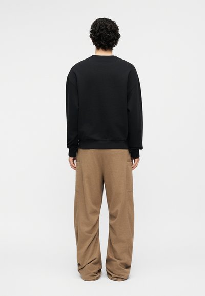 Άτομο με σγουρά μαλλιά, γυρισμένο με την πλάτη, φορά μαύρο μακρυμάνικο πουκάμισο και φαρδιά καφέ παντελόνια, με φόντο απλό λευκό.