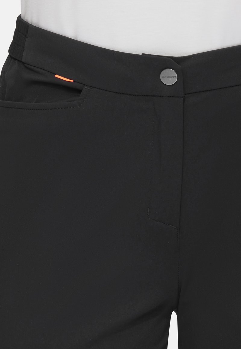 Czarne spodnie z zapięciem na guzik z przodu, bocznymi kieszeniami i małym pomarańczowym detalem z tkaniny w okolicy pasa, noszone z białą bluzką.