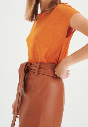 Frau trägt ein ärmelloses orangenes Oberteil, das in einen hoch taillierten braunen Lederrock mit gebundenem Gürtel gesteckt ist, stehend vor einem schlichten hellen Hintergrund.