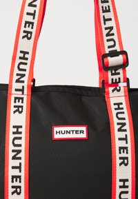 Bolsa tote de nailon negra con asas anchas en rojo y blanco que presentan el texto "HUNTER". Incluye un parche rectangular con el logo y ribete rojo.