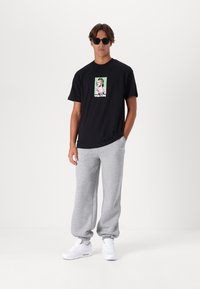 Zwarte katoenen t-shirt met grafische print, grijze fleece sweats en witte sneakers met elastische boorden. Zonnebril maakt de outfit compleet.