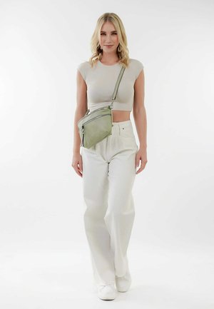 Crossbody taske i lys grøn læder med to lynlåslommer, båret over en tætsiddende beige top og hvide bukser, model stående mod en hvid baggrund.