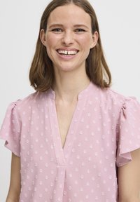 Blusa rosa con scollatura a V e maniche corte a volant. Presenta un ricamo floreale testurizzato su tutto il tessuto. Tessuto leggero, morbida caduta.