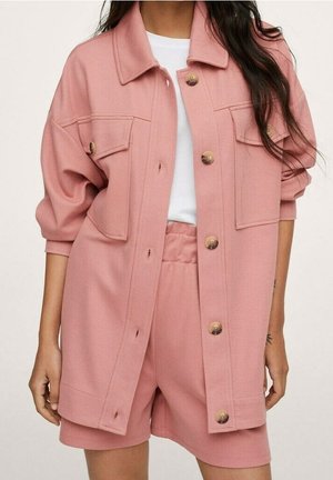 Femme portant une veste rose ample boutonnée avec deux poches poitrine, par-dessus un short taille haute assorti et un haut blanc.