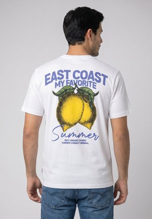 Mann, der ein weißes T-Shirt mit großen gelben Zitronen und dem Text "East Coast My Favorite Summer" auf dem Rücken trägt und vor einem einfarbigen Hintergrund steht.