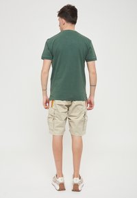 Maglietta in cotone verde, vista posteriore, abbinata a pantaloni cargo beige con tasche laterali e strisce colorate. Scarpe sportive bianche.