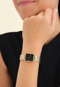 Reloj de metal dorado con una esfera cuadrada negra, con marcadores dorados y una pulsera de eslabones, llevado en una muñeca sobre un fondo de top negro.