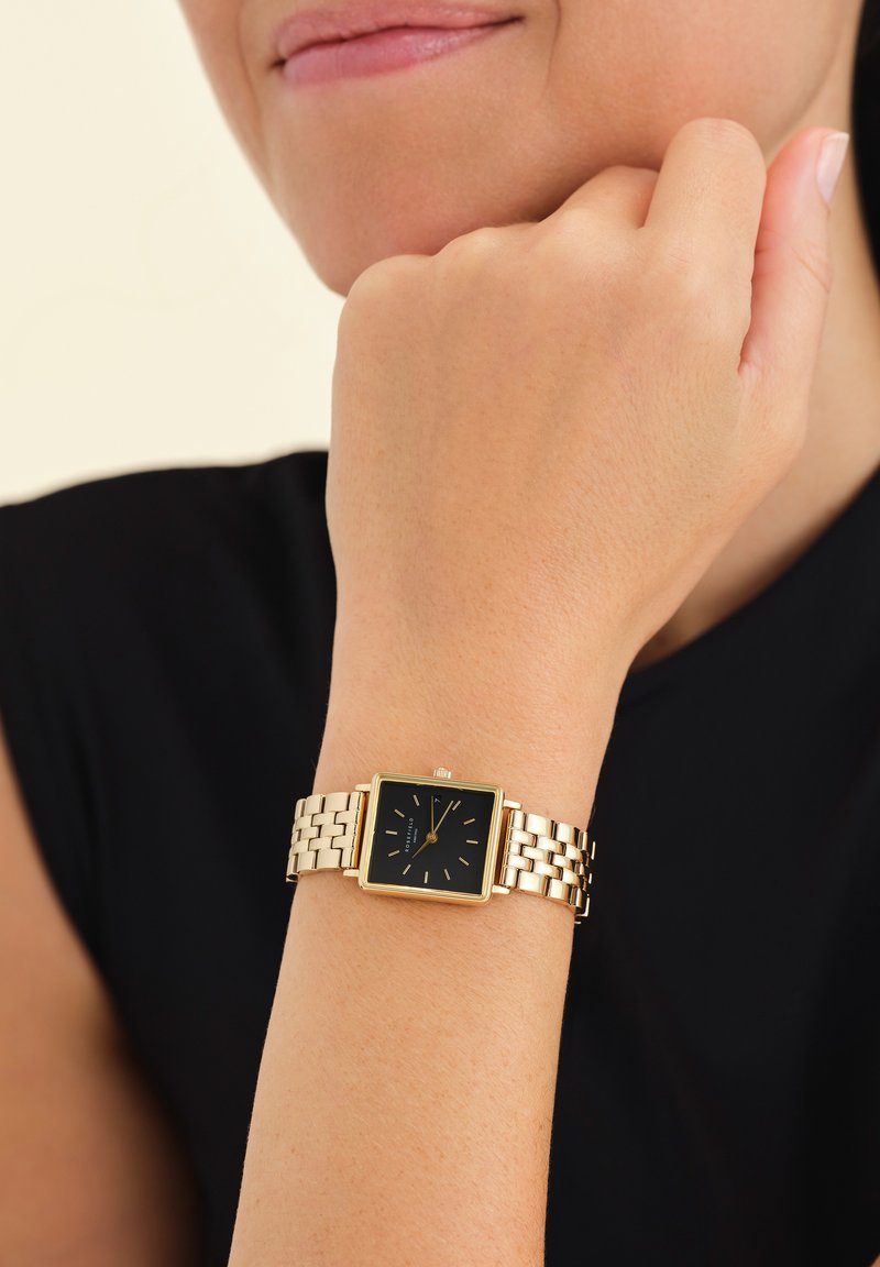 Reloj de metal dorado con una esfera cuadrada negra, con marcadores dorados y una pulsera de eslabones, llevado en una muñeca sobre un fondo de top negro.