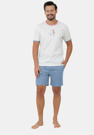 Uśmiechnięty mężczyzna stojący boso, ubrany w biały t-shirt z grafiką biegających postaci i jasnoniebieskie spodenki, ręka w kieszeni.