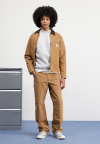 Person with curly hair stands next to gray filing cabinet, wearing tan jacket, matching pants, light gray sweatshirt, and white sneakers.