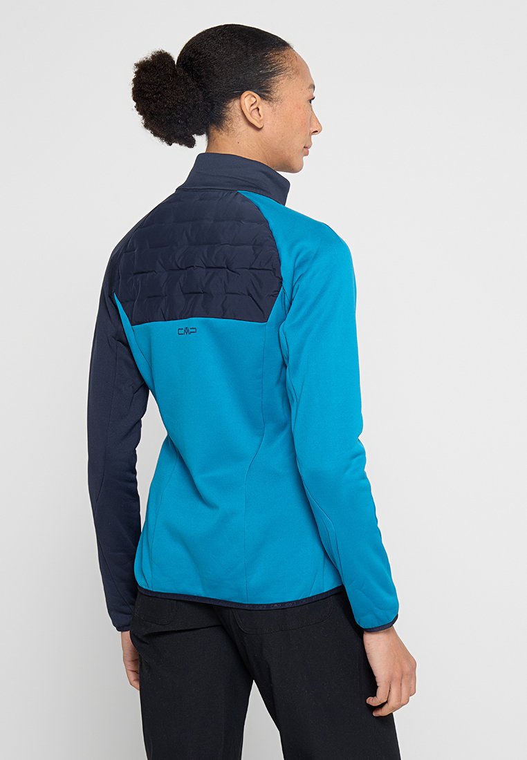 Frau in einer blauen und marineblauen Jacke mit gestepptem oberen Rücken, die seitlich vor einem einfachen hellen Hintergrund steht.