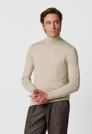 Mann, der einen beigen Rollkragenpullover und dunkelgraue Hose trägt, steht mit verschränkten Händen und schaut seitlich vor weißem Hintergrund.