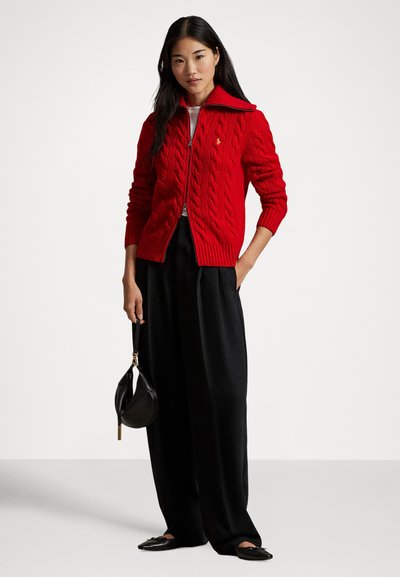 Czerwony kardigan z grubym splotem i wysokim kołnierzem, ozdobiony złotym emblematem. Do tego czarne spodnie o szerokich nogawkach oraz mała czarna torebka.