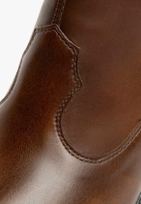 Botín de cuero marrón hasta el tobillo con una textura suave, costuras decorativas y un diseño recortado único en el lateral.
