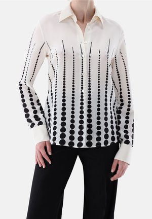 Mujer con blusa blanca de manga larga con botones y puntos negros verticales de tamaños variados, combinada con pantalones negros.