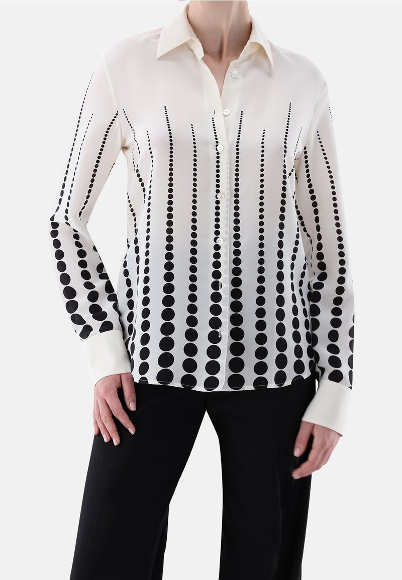 Femme portant un chemisier blanc à manches longues avec des boutons et des points noirs verticaux de tailles variées, associé à un pantalon noir.
