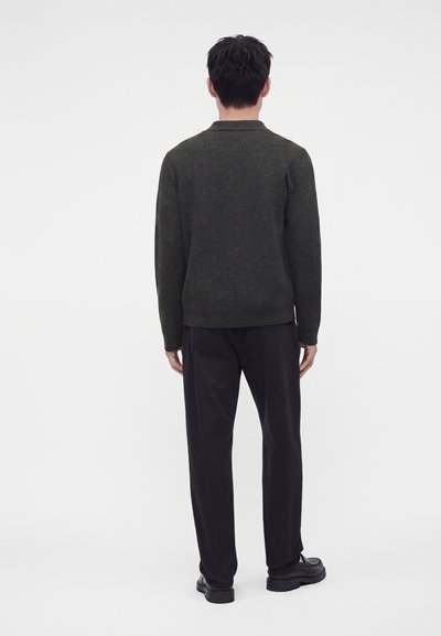 Homme portant un pull vert foncé, un pantalon noir et des chaussures noires, de dos devant un fond blanc uni.