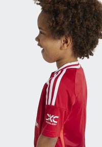 Maillot de sport rouge avec des rayures blanches sur les épaules, à manches courtes, et un logo sur le bras. Texturé doux, coupe athlétique.
