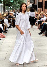 Hvit linantrekk med en løs, kort topp og et langt skjørt med lagdelt design. Lommene er synlige i skjørtet. Flate sko bæres.