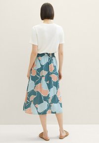 Jupe midi florale en teal, bleu clair et pêche avec un motif texturé et ondulé. Taille élastique, associée à un haut blanc uni et des sandales.