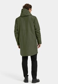 Lång olivgrön vattentät parka med huva, rak passform och sömdetaljer bak, ihop med svarta byxor och ankelskor.