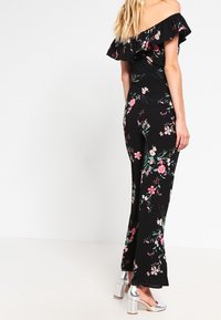 Jumpsuit negro con detalles de volantes sin mangas, que presenta patrones de flores en rosa y verde. Zapatos de tacón cuadrado plateados.