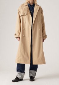 Beżowy dwurzędowy płaszcz przeciwdeszczowy z dużymi guzikami, szerokimi klapami i regulowanymi mankietami, w połączeniu z ciemnym dżinsem i czarnymi skórzanymi butami.