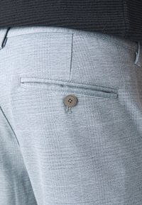 Pantalones gris claro en una tela texturizada que presentan un bolsillo trasero con cierre de botón marrón y un sutil estampado de cuadros.