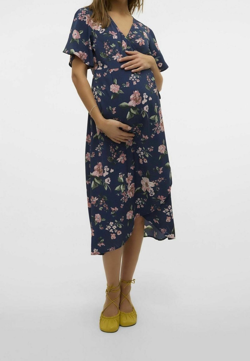 Femme enceinte en robe fleurie bleu marine tenant son ventre, debout avec les pieds croisés, portant des ballerines jaunes à lacets.