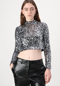 Hopeinen paljettinen pitkähihainen crop-top, jossa on korkea kaula-aukko, yhdistettynä mustiin nahkaisiin korkeavyötäröisiin housuihin, jotka ovat sileäpintaisia ja tiukkaa muotoa.