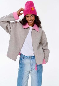 Šedá vlněná krátká bunda s růžovým límcem a manžetami, v kombinaci se světle modrými džíny. Modelka má na sobě jasně růžovou čepici a bílý top.