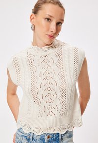 Neulotun ilmanhihaisen puseron väri on off-white ja siinä on korkea kaulus. Puserossa on monimutkaisia pitsikuvioita ja teksturoidut ribbireunat, joita täydentää poikkileikkauksella viimeistelty helma.