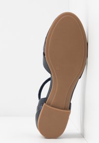 Sandale avec semelle en caoutchouc brun texturé, taille 37, brides supérieures noires, positionnée à la verticale contre un fond blanc en angle.