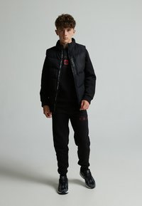 Chaqueta negra acolchada con collar alto, llevada sobre una sudadera negra. Combinada con pantalones de chándal negros y zapatillas con detalles grises. Textura suave.