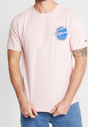 Mann trägt ein hellrosa kurzärmeliges T-Shirt mit einem blauen, runden Logo auf der linken Brustseite und einem kleinen Logo auf dem linken Ärmel.