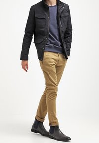 Muž kráčející v černé bundě, modrém svetru, béžových kalhotách a černých Chelsea botách na bílém pozadí.