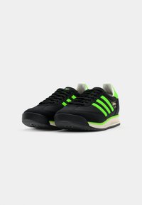 Black sneakers with neon green accents, featuring three stripes design, textured fabric upper, white interior, and black rubber sole.
