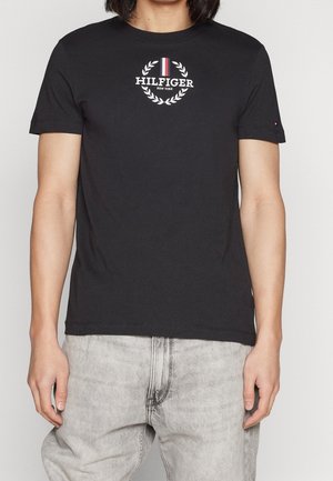 Schwarzes Baumwoll-T-Shirt mit Rundhalsausschnitt, das ein weißes rundes Logo und einen Lorbeerkranz auf der Brust zeigt, kurze Ärmel und eine lockere Passform hat.