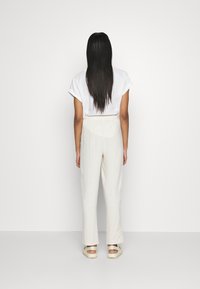 Vit topp med kort ärm som matchas med ljusbeige byxor med elastisk midja. Modell som bär släta, platta, ljusa skor. Enfärgad bakgrund.