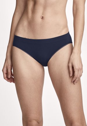 Frau trägt schlichte dunkelblaue Bikini-Unterwäsche, gezeigt vom mittleren Oberkörper bis zu den Oberseiten der Oberschenkel, mit entspannt an den Seiten hängenden Armen.