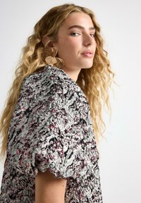 Teksturoitu metallipaita hopean, mustan ja vaaleanpunaisen sävyissä. Omaavat puffaushihat, pyöreän kaula-aukon ja monimutkaisia kuvioita. Suuret metallikorvakorut.