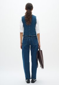 Denim jumpsuit met een getailleerd vesttop en losse broeken in medium blauw, met achterzakken. Model houdt een donkerbruine leren draagtas vast.