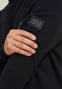 Marineblauw trui van textuurgebreide stof, met een zwart leren logo-patch op de mouw. Close-up van een hand die de stof vasthoudt.