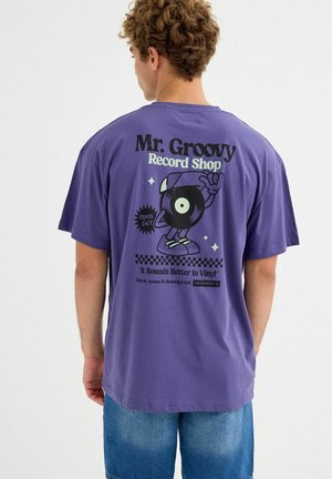 Hombre con cabello rizado que lleva una camiseta morada con un personaje de disco de vinilo caricaturesco y texto que promociona "Mr. Groovy Record Shop" en la parte trasera.