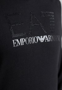 Crna pamučna dukserica s metalik srebrnim logotipima "EA" i "Emporio Armani" uz dodatak rhinestone akcenta, izrađena od glatkog teksturiranog materijala.