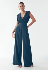 Teal ühes tükis, millel on sügav V-kaelus, volditud varrukad, laiad püksijalad, tekstureeritud kangas ja sujuv disain, mida täiendab kuldne kott.