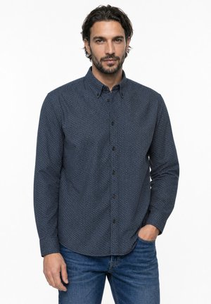 Homme aux cheveux courts et foncés, barbe, portant une chemise sombre à motifs boutonnée et un jean bleu, debout avec une main dans la poche.